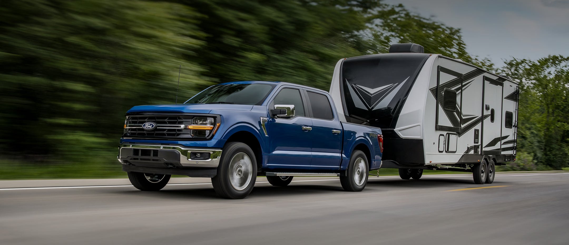 Blue Ford F-150 towing a large trailer on a highway with trees in the background.