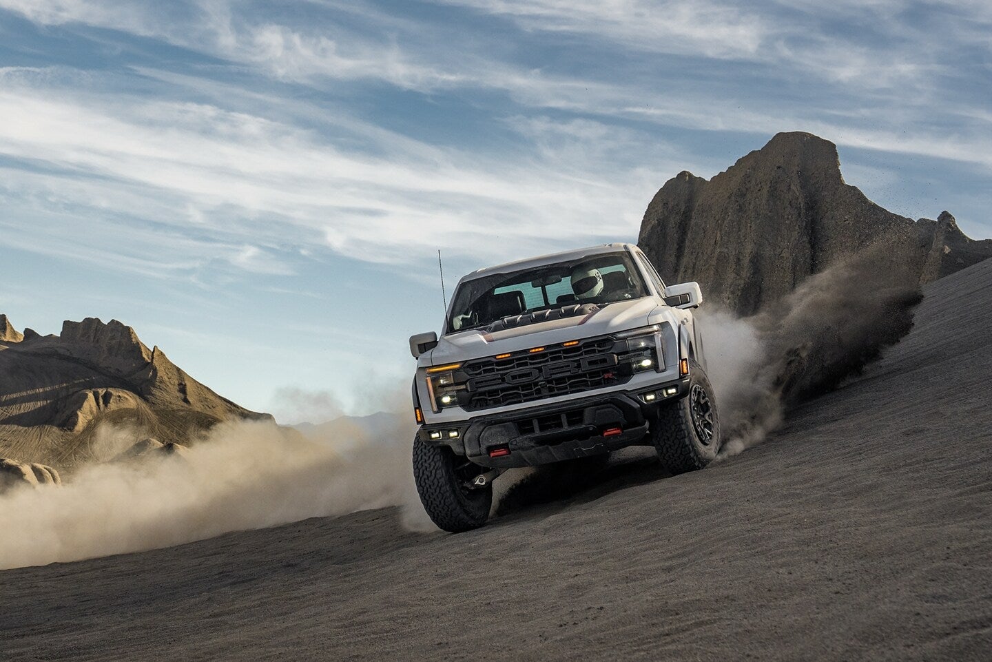White Ford F-150 Raptor kicking up dust while driving on rugged terrain against a backdrop of mountains and blue sky