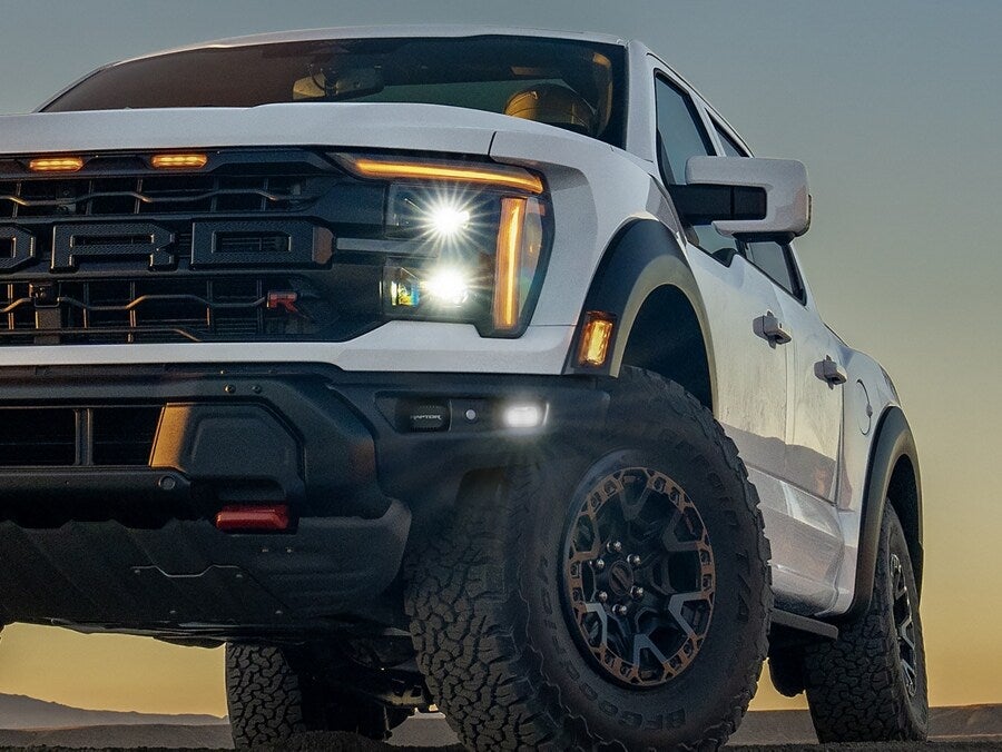 Close-up of a white Ford F-150 Raptor with rugged tires and illuminated headlights.